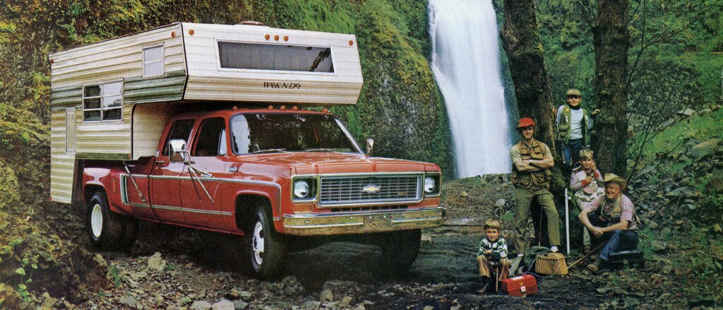 Una foto del folleto de camionetas Chevy del 1973 original con una pickup de la serie C/K roja con casilla en la caja, estacionada en un área boscosa cerca de una cascada. Al lado de la camioneta se ve a un hombre y cuatro muchachos de diferentes edades sentados y de pie.