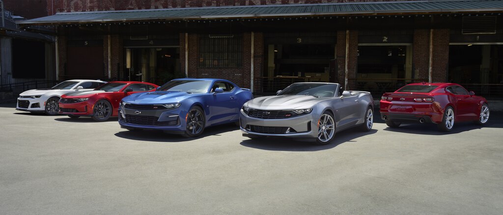 Un grupo de cinco Camaro estacionados en un muelle de carga: Coupes en color blanco, rojo y azul y un convertible en color plata.