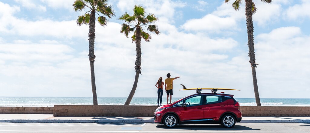 Un Chevrolet Bolt EV rojo con una tabla de surf sujeta con sogas a la parrilla portaequipajes en el techo frente a tres palmeras, con la playa y el océano más allá de los árboles. Dos personas de pie en una pared baja detrás del Bolt EV, señalando al agua.