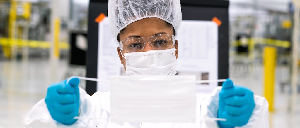 Una persona usando mascarilla, redecilla y guantes muestra una mascarilla sosteniéndola de los elásticos en ambos lados.