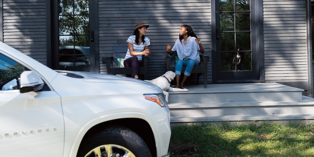 dos mujeres se sientan y conversan sobre el porche de una cabaña, con una Chevrolet Trverse color Blanco en el fondo.