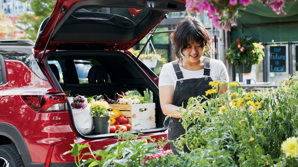 La chef Ji Hye Kim sonríe mientras observa un despliegue de flores amarillas en un mercado agrícola. Detrás de ella, una Chevy Trailblazer roja estacionada con el maletero trasero abierto con el área de carga llena de frutas, verduras y flores.