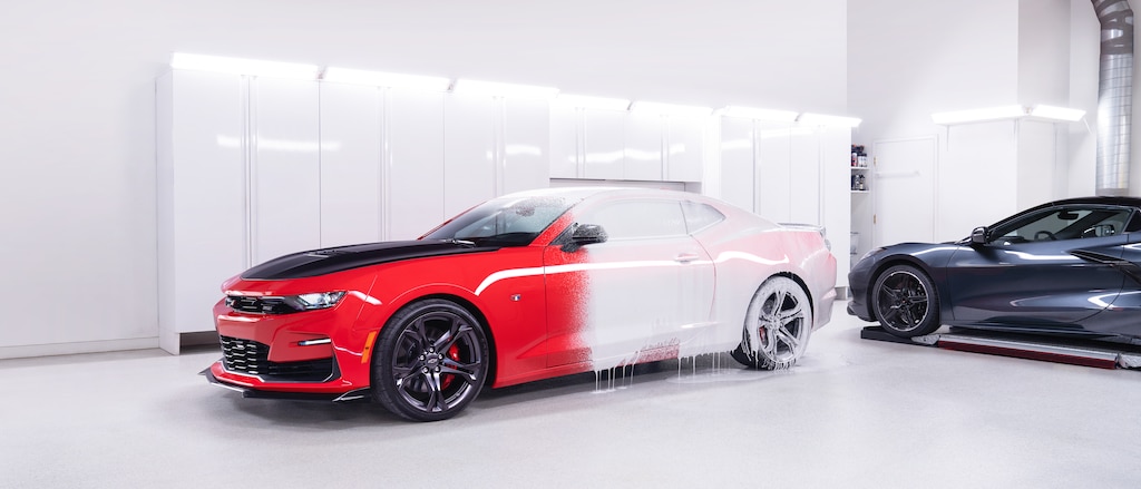 Un Chevy Camaro color Rojo Brillante cubierto parcialmente de espuma blanca en el inmaculado interior blanco de la tienda Esoteric Fine Auto Finishing al lado de un Corvette 2020 con motor central en color Gris Oscuro.