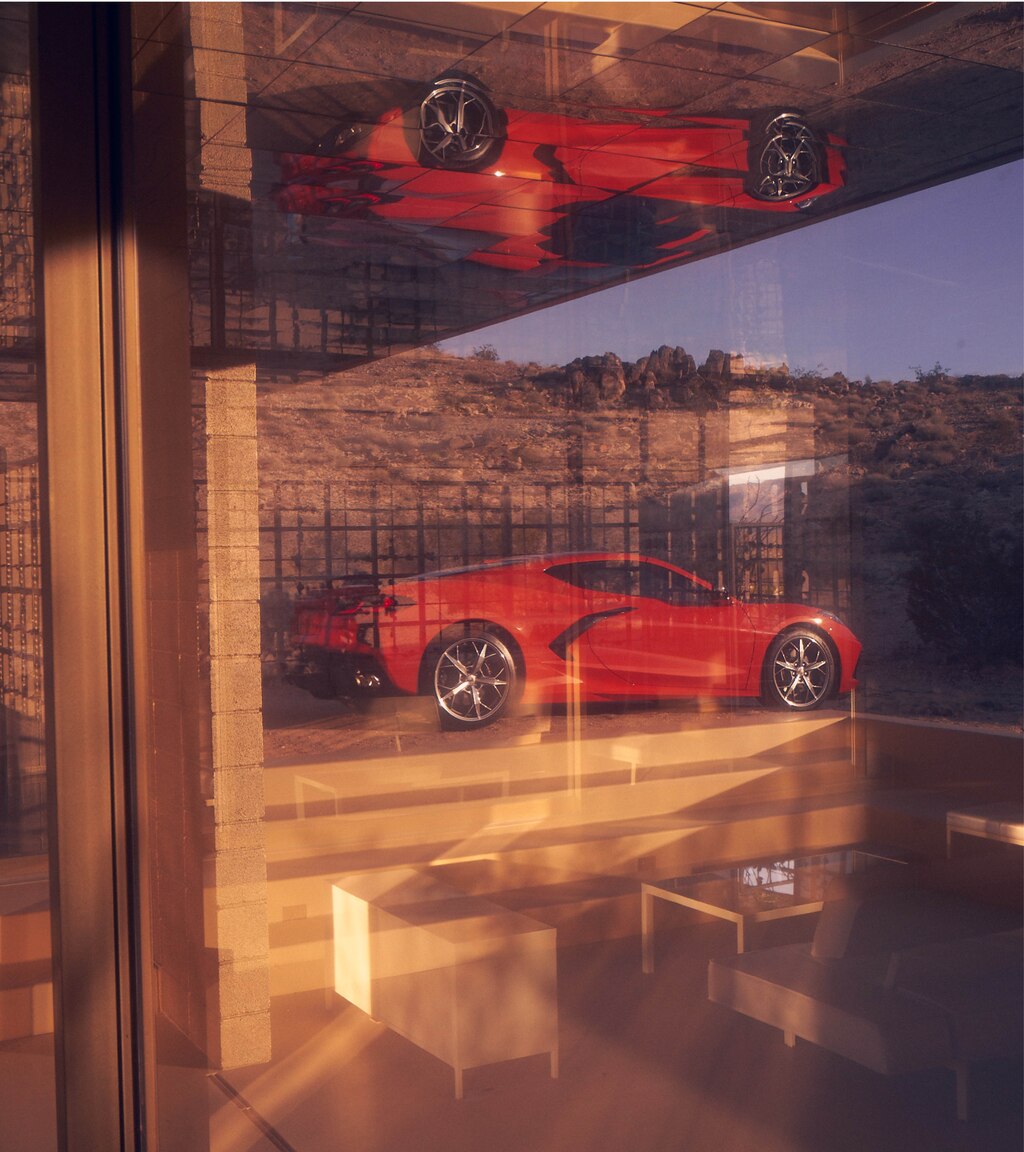 Un coupe Corvette 2020 en Rojo Antorcha en un paisaje desértico, visto a través de una ventana. En el vidrio se ve el reflejo del auto.