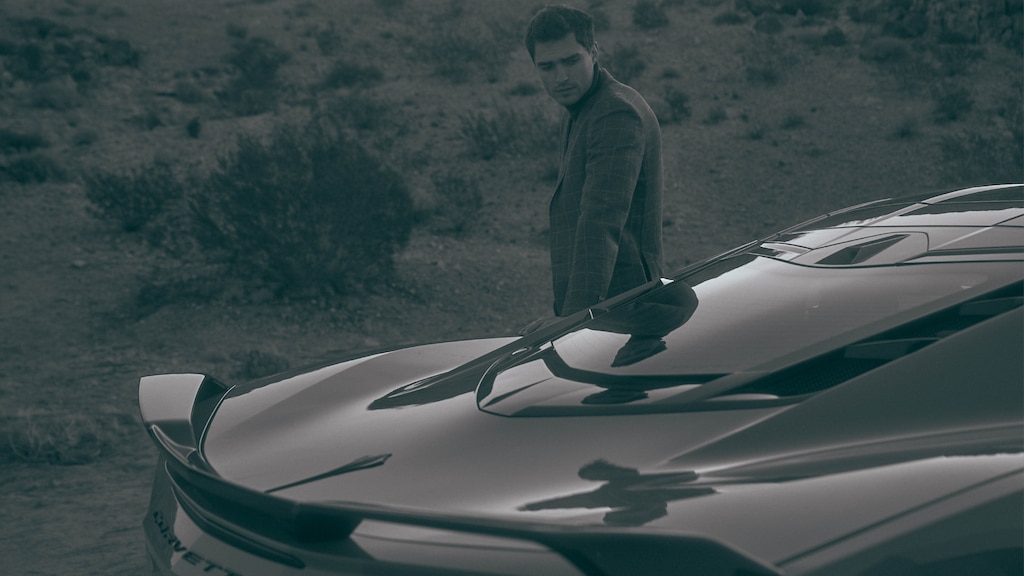 Un hombre del lado opuesto de un Corvette 2020 mira la parte de atrás del auto, visto desde un ángulo ligeramente sobre el auto para mostrar el parabrisas trasero y parte trasera.