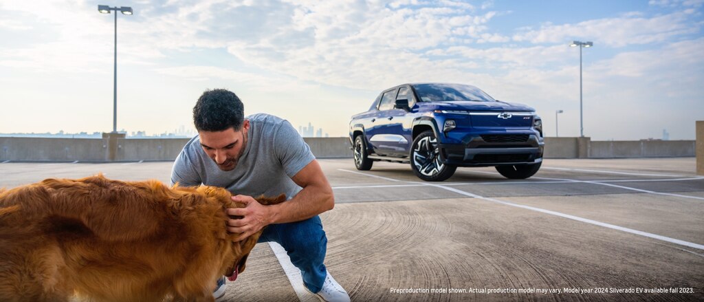 Una persona se arrodilla para acariciar a un golden retriever en un estacionamiento mientras una Silverado EV azul se encuentra en el fondo.