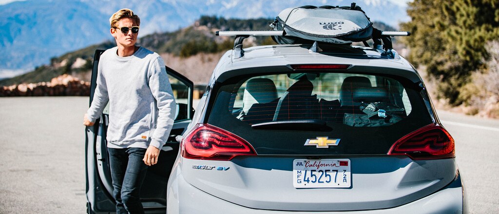 Kevin Schulz, con gafas de sol, sale por la puerta del conductor del Bolt EV con una tabla de surf sujeta a la parrilla portaequipajes en el techo.