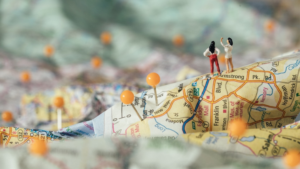 Una imagen tipo maqueta de un mapa de papel 3D con pines amarillos adheridos y dos muñequitos de pie en una cresta.
