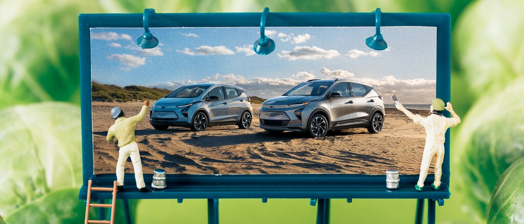 Una imagen tipo maqueta de una valla miniatura mostrando el Bolt EV y la Bolt EUV, con dos muñequitos pintándola, rodeada de coles de Bruselas.