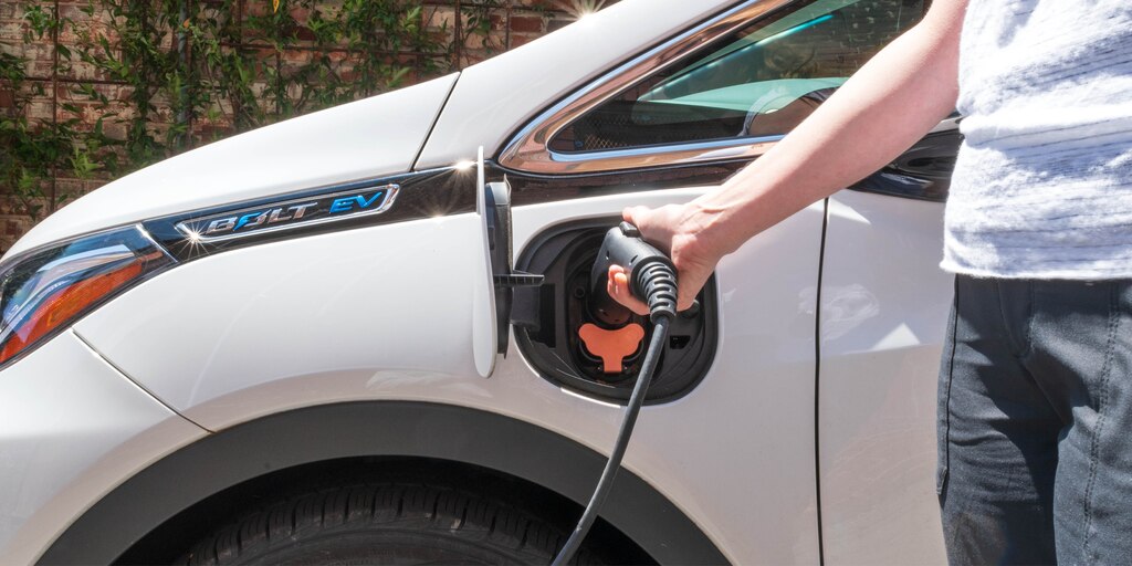 Se ve el brazo de una mujer enchufando un cable de carga en uno de los lados de un Bolt EV blanco.