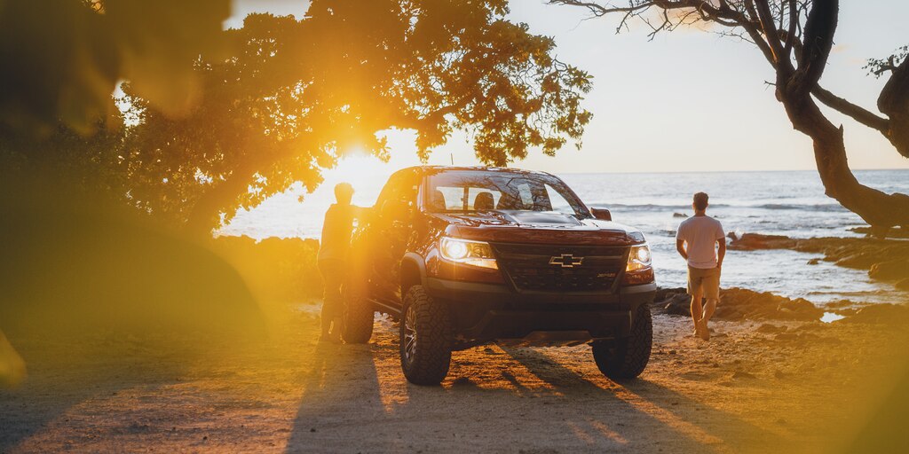 Una Chevrolet Colorado ZR2 roja estacionada en la playa al atardecer. Un hombre descansa apoyado en la camioneta mientras otro camina hacia el agua.