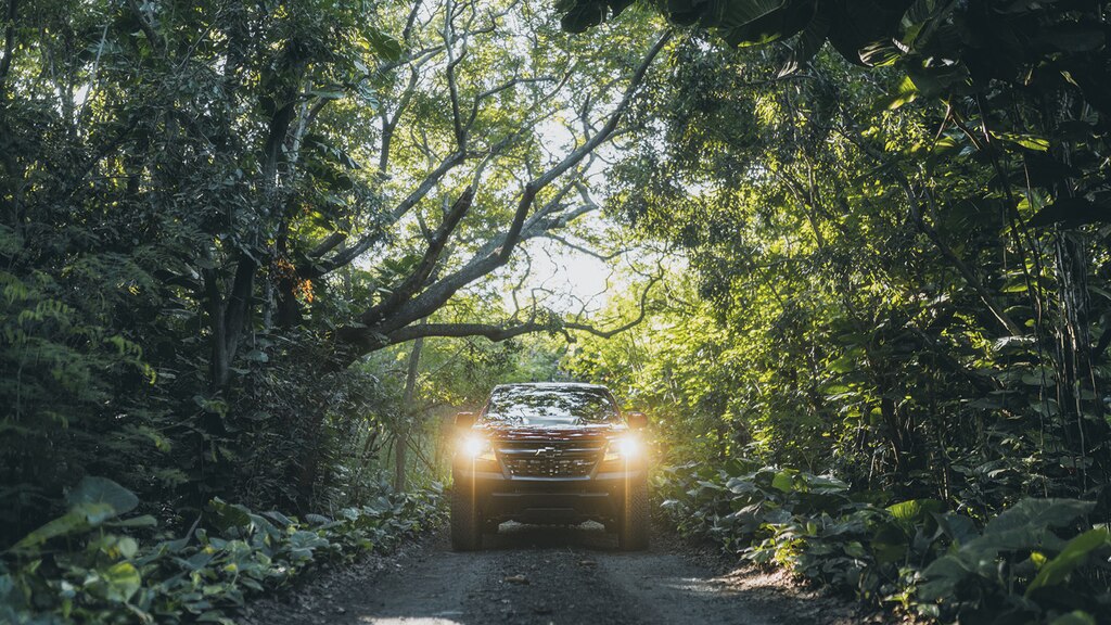 Vista delantera de una Chevrolet Colorado ZR2 con los faros delanteros encendidos andando por un camino con una vegetación densa en ambos costados.