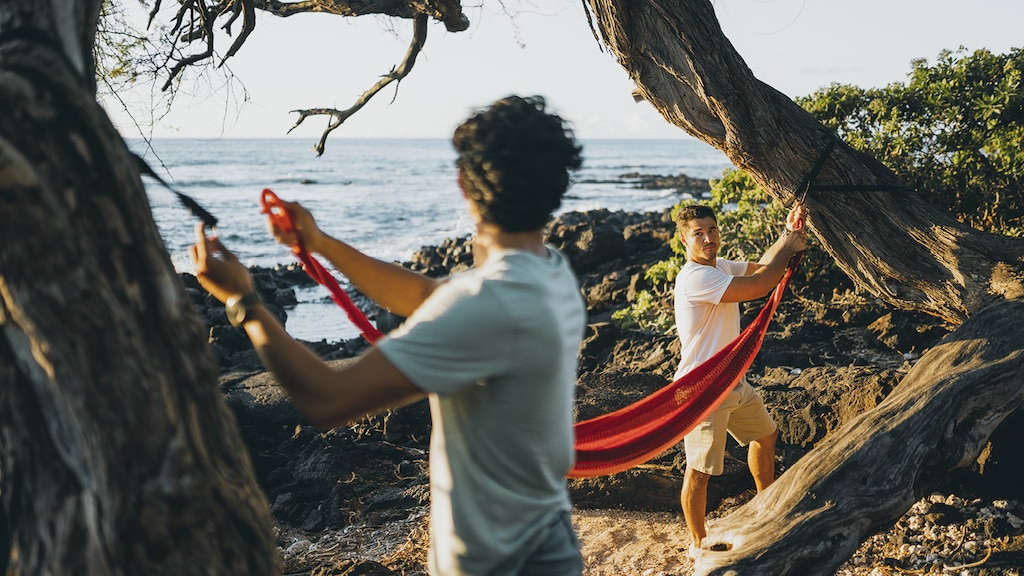 Dos hombres colgando una hamaca roja entre dos árboles en la playa y con el agua de fondo.