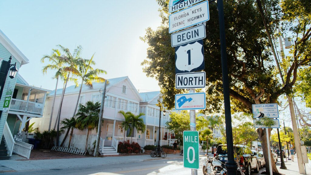 Un poste indicador con un cartel que dice "Florida Keys Scenic Highway. Begin Highway 1 North. Mile 0". En el fondo se ven edificios de colores pastel y palmeras.