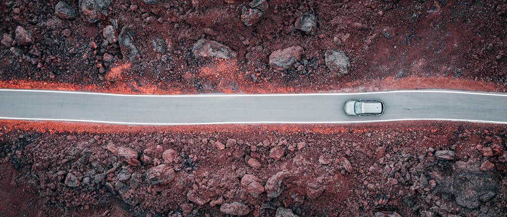 Una toma aérea desde un dron de una Chevrolet Blazer gris transitando un camino negro a través de un campo de rocas de lava.