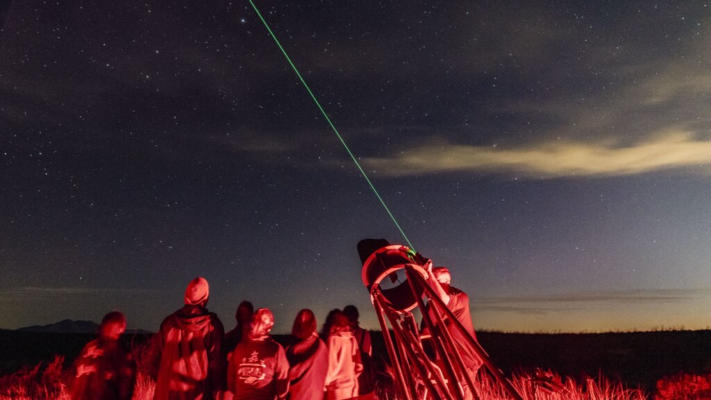 Un grupo de personas se reúnen alrededor de un telescopio que tiene una guía láser señalando hacia el cielo que se va oscureciendo. Al grupo lo ilumina una luz roja para no interferir con la visión nocturna.