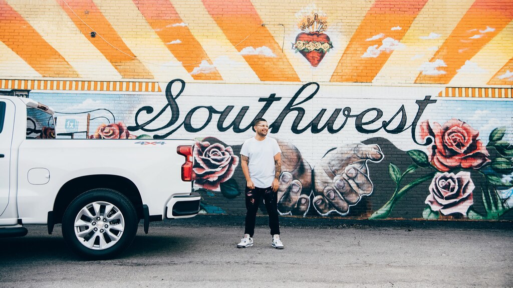 Freddy Diaz de pie detrás de una Silverado blanca, frente a uno de sus coloridos murales con la palabra "Southwest", un par de manos y rosas.