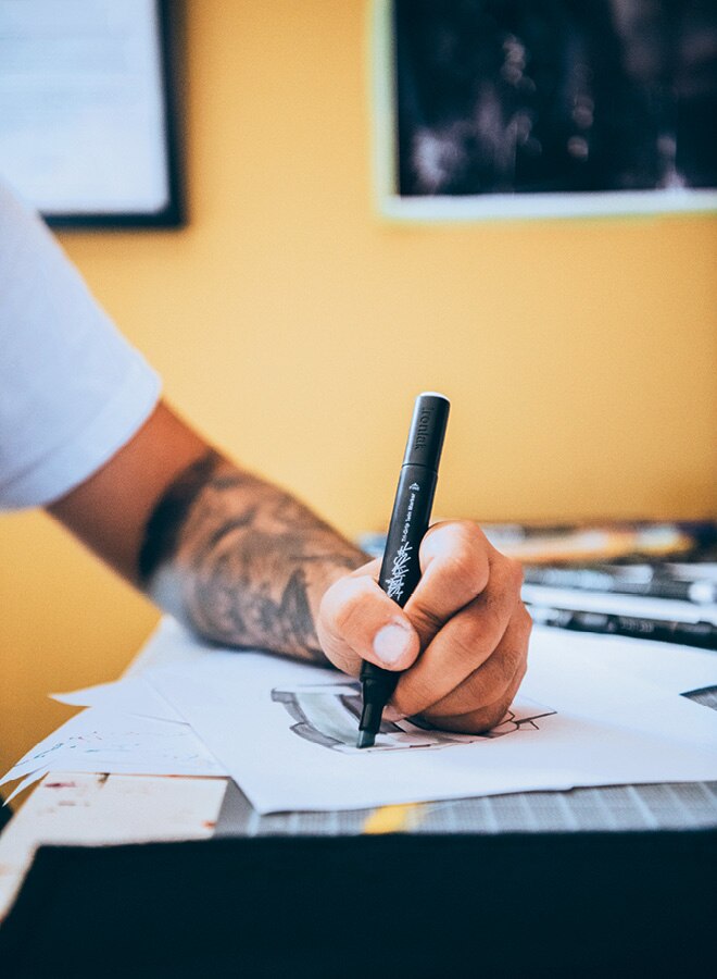 El brazo tatuado y la mano de Freddy se pueden ver mientras dibuja en papel sobre un tablero de dibujo técnico.