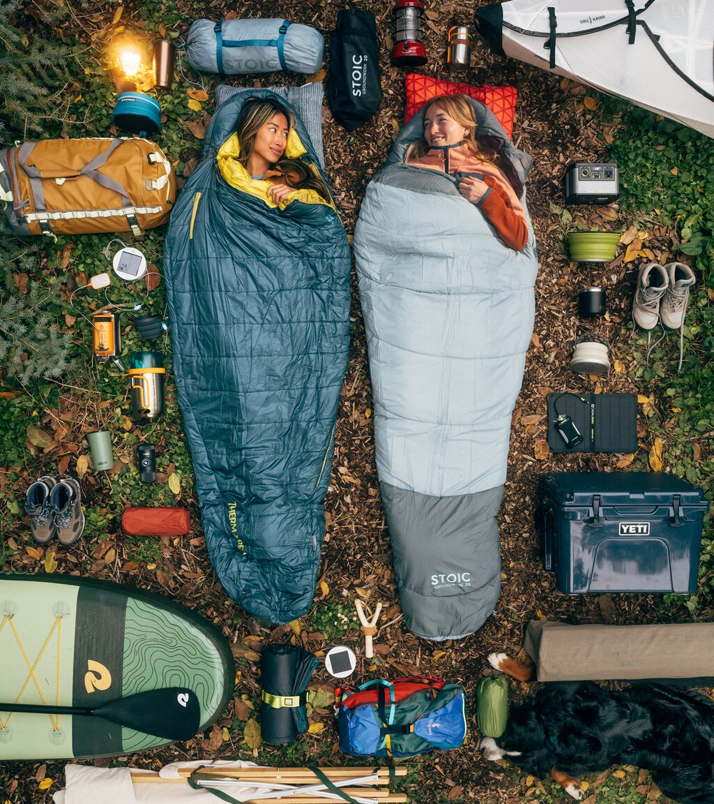 Vistas desde arriba, dos mujeres están acostadas en el suelo en un campamento, en bolsas de dormir rodeadas por una gran variedad de artículos de campamento.