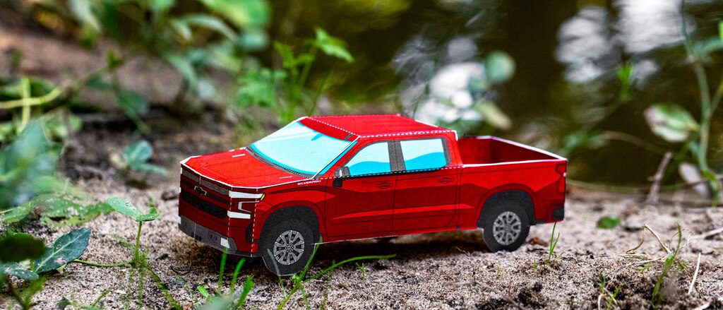 Un modelo en papel miniatura de una Silverado RST 2020 diesel en Rojo Cajún sobre terreno descubierto con grama y hojas en el fondo.