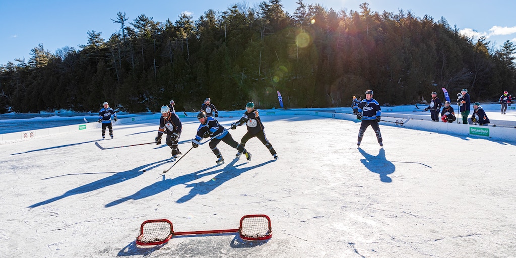 Un grupo de personas jugando al hockey sobre un estanque congelado, con un arco bajo al frente y árboles de fondo.