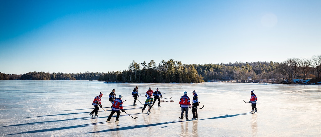 Un grupo de personas jugando al hockey sobre un estanque congelado, con un fondo arbolado y un cielo azul y despejado.