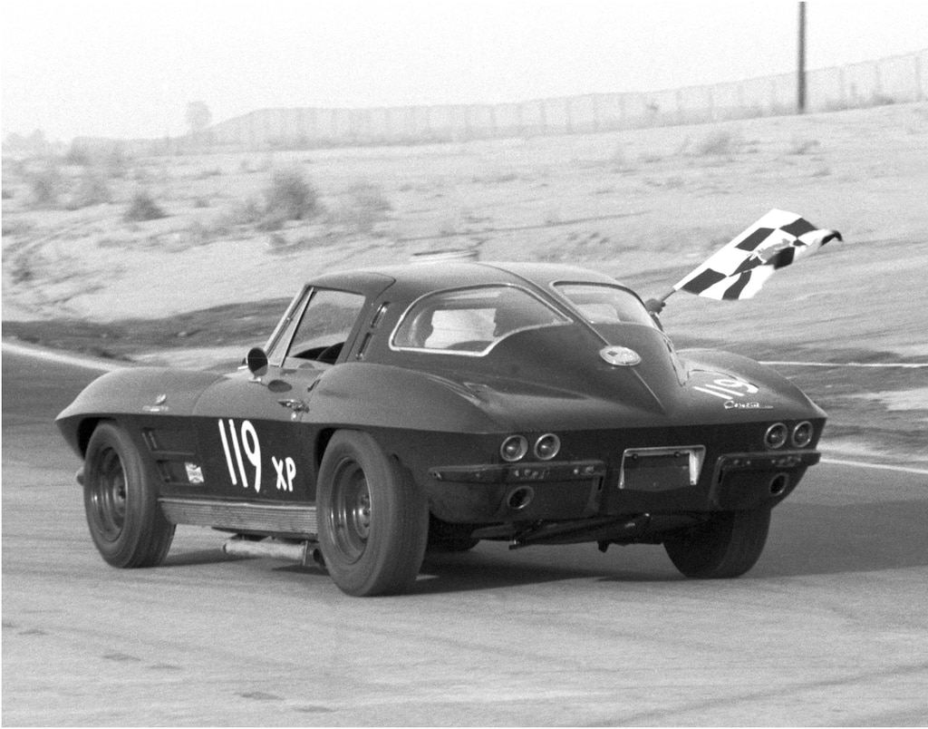 Una foto histórica de un Corvette Sting Ray de ventana dividida con el número 119 pintado en el lateral y una bandera a cuadros blanca y negra ondeando por la ventanilla del pasajero circulando por un circuito de carretera.