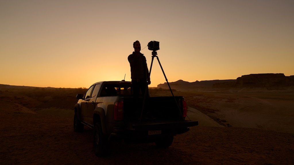 Hombre parado en la caja de una pickup Colorado ZR2 con una cámara de gran formato en un trípode, con el sol poniéndose de fondo.
