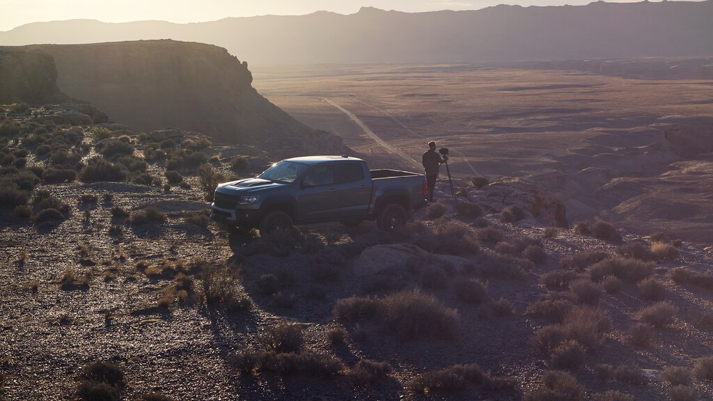 Una Chevy Colorado ZR2 Bison gris en el borde de una pendiente pronunciada cubierta de vegetación al atardecer. Hombre parado detrás de la camioneta usando una cámara de gran formato montada en un trípode.
