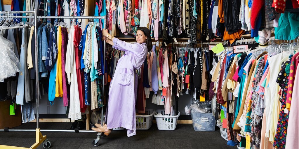 Beth Morgan parada sobre un rack de ropa portátil lleno de vestuarios frente a una pared de racks llenos de principio a fin de vestuarios.