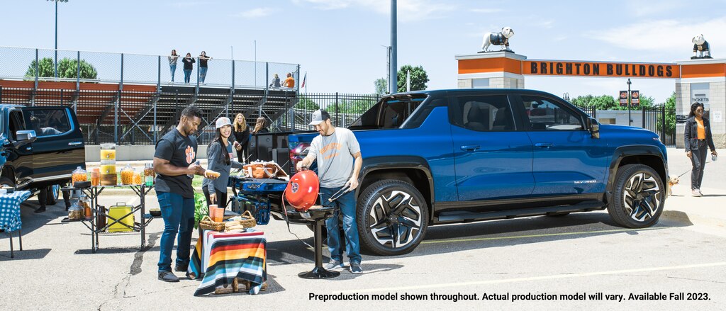 Un grupo de personas se reúnen alrededor de la puerta trasera de una Silverado EV en el estacionamiento de un estadio con una parrilla portátil y una mesa portátil repletas de comida y bebida, además de otros artículos en la puerta trasera de la Silverado.