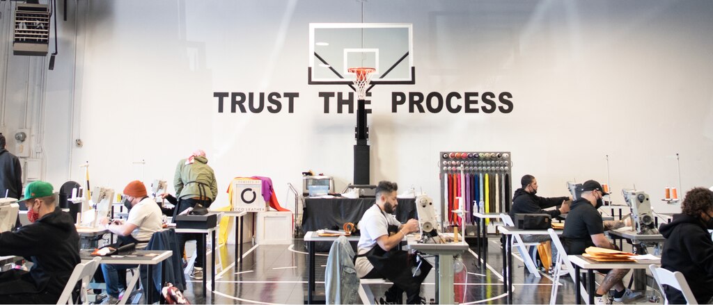 The Sneaker School se instaló en el gimnasio de una escuela, con casi una docena de personas sentadas en mesas con máquinas de coser. Un letrero detrás de una canasta de básquetbol que dice <em>Trust the Process</em> (Confía en el proceso).