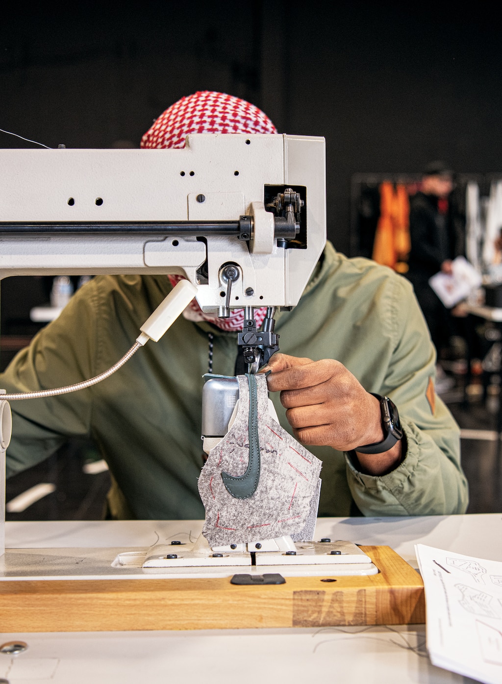 Una persona usando una bandana a cuadros rojos y blancos y una camisa verde trabaja en una máquina de coser industrial, su rostro queda oculto tras la máquina. En la máquina hay una pieza de tela blanca con un logotipo de Nike verde.