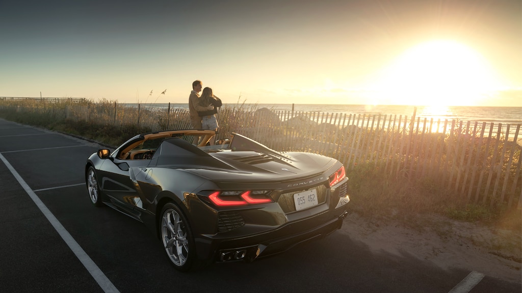 Dos personas viendo el atardecer de pie junto a un Corvette Convertible 2020 en Bronce Zeus Metálico estacionado junto a un área costera en Carolina del Sur.
