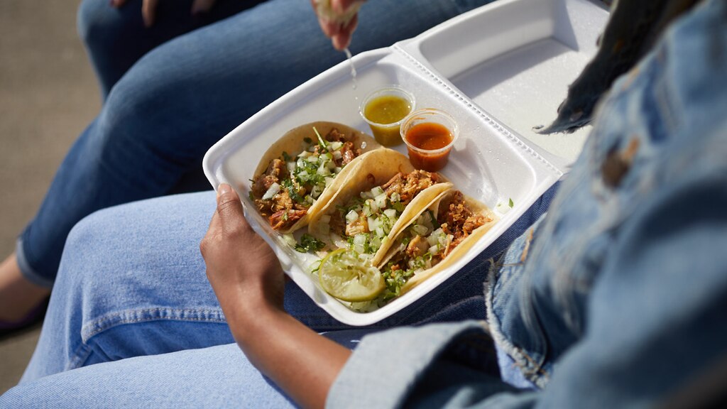 Tres tacos y salsas adentro de una caja grande de comida para llevar, que está en el regazo de una persona usando jeans y chaqueta de jean.