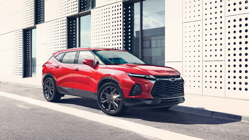 La parte delantera y el lado del acompañante de una SUV Blazer RS en color Rojo estacionada al lado de un edificio de diseño contemporáneo.