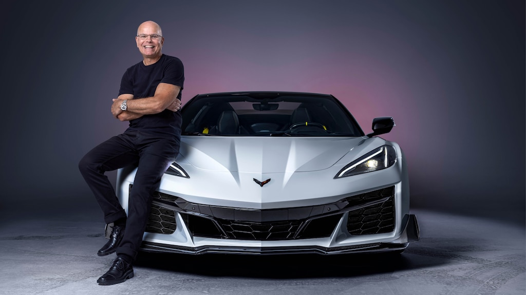Jordan Lee, con los brazos cruzados y sonriendo a la cámara, se sienta en la parte delantera derecha de un Corvette Z06 Blanco que está de frente a la cámara.