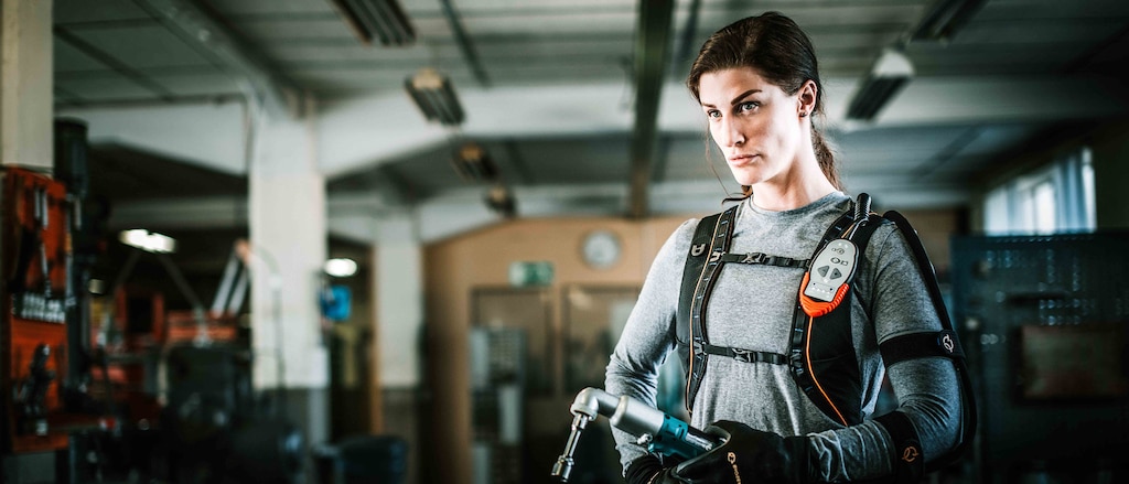 Una mujer usando guantes y exoesqueleto de alta tecnología para aumentar su fuerza, de pie en una planta industrial con una llave de impacto en la mano.