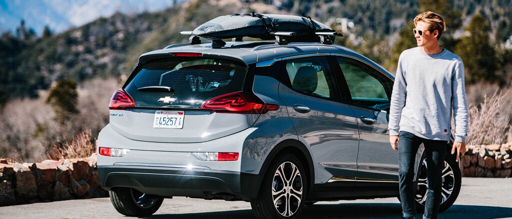 El surfista profesional Kevin Schulz se aleja de un Bolt EV estacionado al costado de un camino de montaña con una tabla de surf y un snowboard sujetos a la parrilla portaequipajes en el techo.
