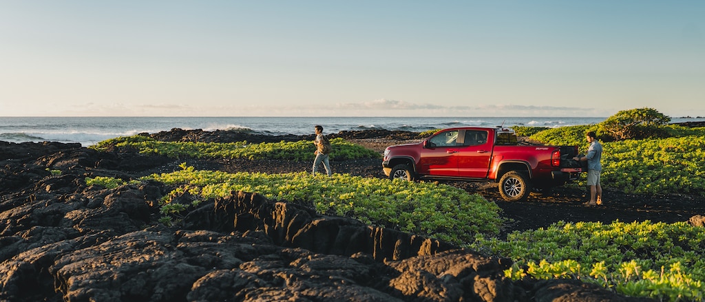 Dos personas de pie cerca de una Chevrolet Colorado ZR2 roja estacionada en un área rocosa con arbustos bajos junto al mar en Hawái.