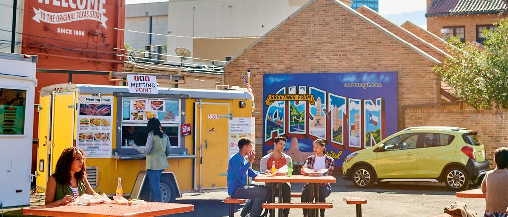 Varias personas comiendo en mesas en la zona de camiones de comida de la universidad en Austin, Texas, con un Chevy Spark 2018 amarillo en el fondo.