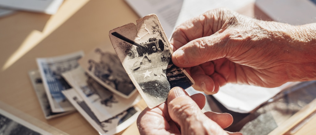 Manos de George Patton Waters sosteniendo una fotografía vieja de un tanque, junto a otras fotos antiguas en la mesa que está debajo.