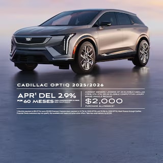 Imagen de un SUV eléctrico Cadillac OPTIQ plateado estacionado sobre una superficie lisa y reflectante al anochecer, visto desde una perspectiva frontal de tres cuartos. El texto grande dice "Modelos Cadillac OPTIQ 2025/2026", "0.9% APR durante 72 meses," y "$2,000 de descuento en la compra," con detalles de la oferta en letra pequeña en la parte inferior.